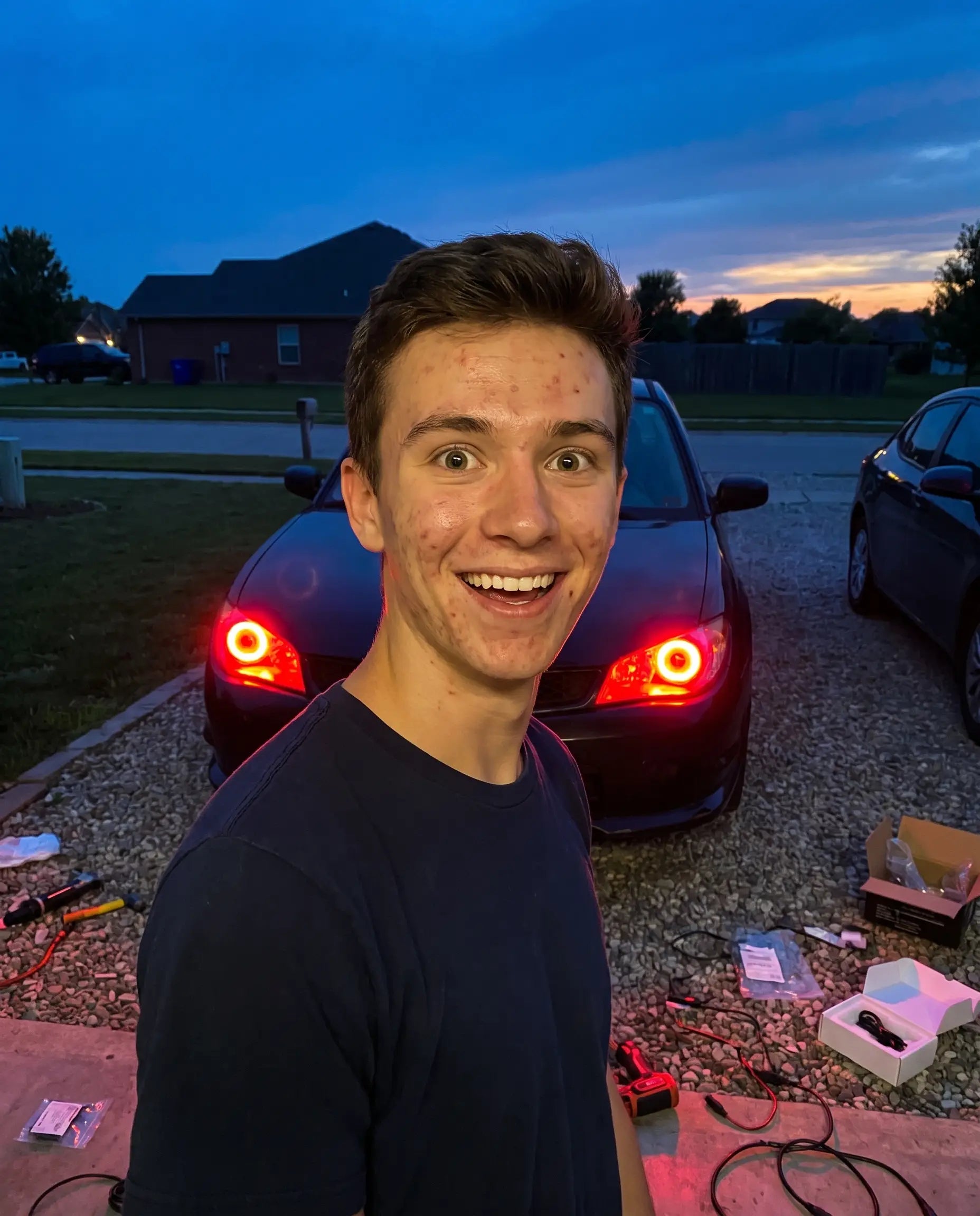 Car owner smiling at newly installed Nyxora Demon-Eye LED headlights glowing red in driveway at dusk