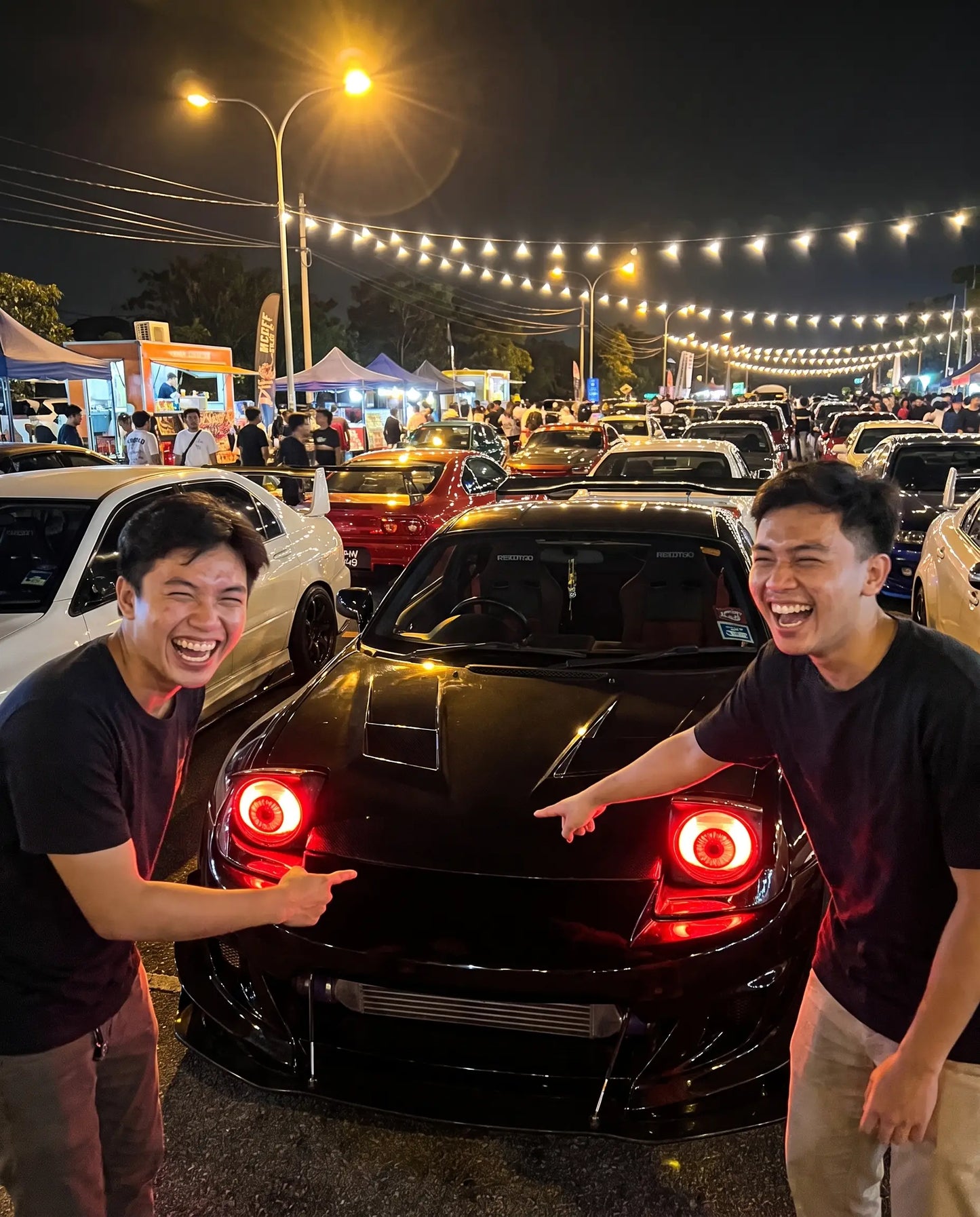 Two friends at a night car meet pointing at a car's glowing red Nyxora Demon-Eye LED headlights